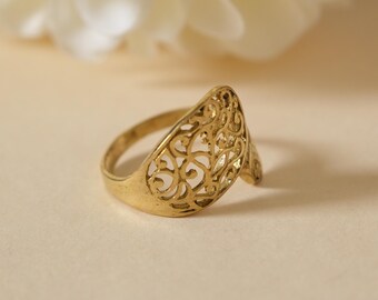 May include: A gold-colored ring with an openwork design. The ring features an intricate, swirling pattern on the face, with a smooth band. The ring is set against a soft, neutral background, with a blurred white flower in the background.