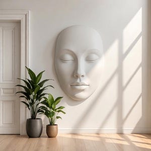 Puede incluir: Una gran escultura de pared blanca de forma ovalada que representa un rostro humano sereno con los ojos cerrados, una nariz definida y labios neutros. La escultura está montada en una pared lisa junto a dos plantas verdes en macetas gris y beige.