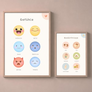 May include: Two framed prints with light wood frames. The larger print displays colorful emoji-style faces with German words for emotions. The smaller print shows illustrations with German words for needs. Both prints have a soft, neutral background.