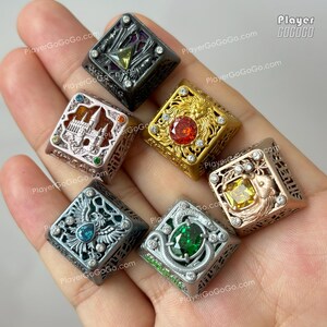 May include: Six square metal keycaps featuring intricate relief designs and embedded gemstones. The designs include a castle, a lion, a bird, a snake, and geometric patterns. Each keycap is finished in metallic tones of silver, gold, and dark grey.