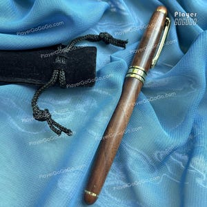 May include: A wooden pen with gold-colored accents and a gold clip. The pen is dark brown and has a smooth finish. A black pen sleeve with a drawstring is next to the pen. The background is a blue fabric.