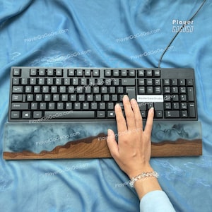 May include: Black computer keyboard with white lettering and a matching wrist rest. The wrist rest features a wood and resin design with a blue and brown colour scheme. The keyboard has the text "MasterGearStudio" on it.