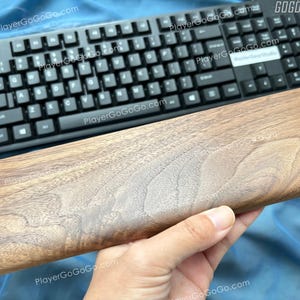 以下が含まれることがあります： 白い文字が入った黒いコンピュータキーボードと木製のリストレスト。 リストレストは、木目模様の明るい茶色です。 キーボードには、右上隅に「GOGOGO」というテキストがあります。