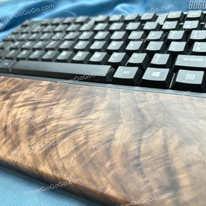 Op de afbeelding: Een zwart computertoetsenbord met witte letters en een houten polssteun. De polssteun heeft een uniek houtnerfpatroon. Het toetsenbord ligt op een blauw oppervlak. De tekst "GOGOGO" is rechtsboven zichtbaar.