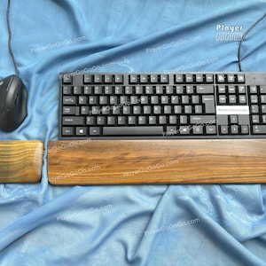 Op de afbeelding: Een zwart computertoetsenbord met bijpassende muis en houten polssteun. De polssteun en muismat zijn gemaakt van lichtbruin hout. Het toetsenbord heeft een label met de tekst "MasterGeekStudio". De items liggen op een blauwe stof.