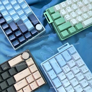 May include: Four mechanical keyboards with different colour keycaps are displayed. The keyboards have various colour schemes, including white, blue, green, and black. The keyboards are arranged on a blue fabric surface. The website PlayerGoGo.com is watermarked on the image.