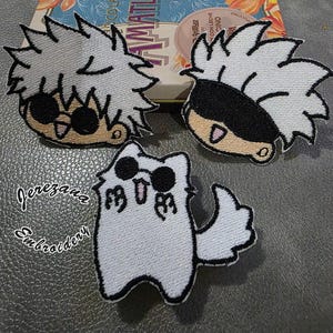 May include: Embroidered patches featuring anime-style characters with spiky hair and a cat-like figure. The patches are in white, black, and tan, with detailed stitching. The background includes a decorative box with floral designs.