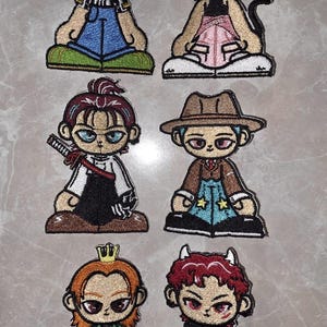 May include: A collection of six embroidered patches featuring stylized cartoon characters. Each patch has a unique design, including characters with green hair, cat ears, a sword, a fedora, a crown, and horns. The patches are in various colors, including blue, pink, and green.