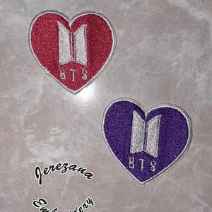 May include: Two heart-shaped embroidered patches, one red and one purple, each featuring the letters "BTS" in white. The patches are on a gray surface. The text "Jerezana Embroidery" is also visible.