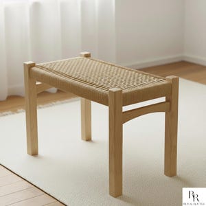 Puede incluir: Un banco de madera de color claro con un asiento tejido. El banco tiene cuatro patas robustas y un asiento rectangular hecho de material tejido. La madera tiene un acabado natural y el banco está colocado sobre una alfombra de color claro.
