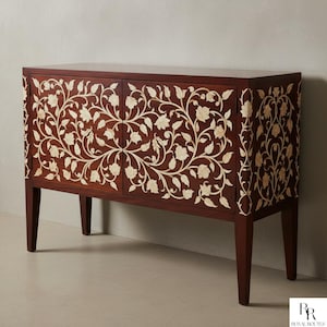 Puede incluir: Un mueble de madera marrón oscuro con dos puertas, con un intrincado diseño floral en color crema. El mueble tiene cuatro patas y una forma rectangular, adecuado para el almacenamiento.