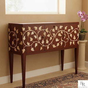 May include: A dark brown wooden cabinet with three doors and four legs. The cabinet is decorated with a cream-coloured leaf and vine design. The cabinet is in front of a window and a decorative rug.