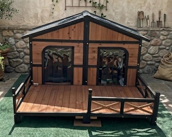 May include: A wooden dog house with a deck and a black roof. The dog house has two entrances with clear plastic flaps. The structure is made of brown wood with black trim and a small railing around the deck.