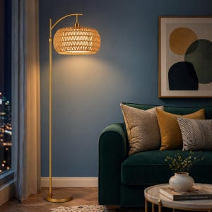 May include: A gold floor lamp with a woven rattan shade. The lamp has a curved arm and a round base. The shade is cylindrical and emits a warm glow. The lamp is next to a green sofa and a painting.