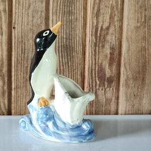 May include: A ceramic figurine of a penguin standing on a blue and white wave base. The penguin has a black back, white chest, and yellow beak. Next to the penguin is a white, shell-shaped open container with a scalloped edge and a glossy finish.