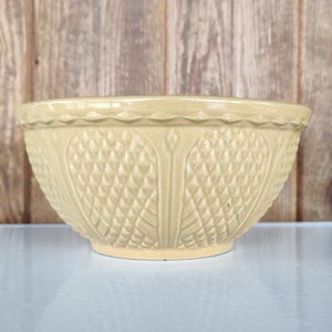 May include: A beige ceramic mixing bowl with a textured diamond pattern and a decorative rim. The bowl is set against a wooden background.