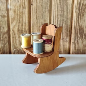 May include: A small wooden rocking chair holds five spools of thread in various colors: yellow, cream, teal, and burgundy. The chair is made of light brown wood and sits on a white surface against a wooden plank background.