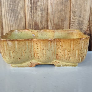 May include: A yellow ceramic planter with a speckled brown glaze. The planter has a rectangular shape with three rounded sections.