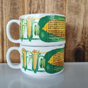 May include: Two white ceramic mugs stacked on top of each other. The mugs have a green and yellow corn design with a recipe for corn soup printed on the side. The recipe reads: 1. Cut kernels from corn scraping as possible. Boil enough chicken stock with a cup of water, salt. Strain and reserve broth. 2. To kernels, add 1/4 cup minced celery, 1/4 cup dry mustard, 1 tsp salt, 1/2 tsp pepper. Sauté and add to broth. 3. Add 1 Tbsp lemon juice.