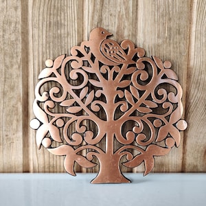 May include: A circular copper-colored metal trivet featuring an intricate openwork design of a tree with swirling branches and leaves, topped with a small bird perched at the center. The metallic surface has a warm, reflective finish.