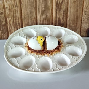 May include: A white ceramic egg serving platter with a textured honeycomb pattern featuring a central decorative element of a yellow chick emerging from a cracked white eggshell nestled in a brown textured nest-like base.