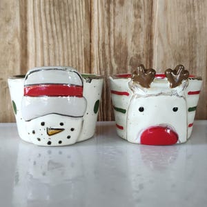 May include: Two ceramic bowls featuring holiday designs. The left bowl is white with green dots and a snowman face wearing a red and white hat. The right bowl is white with red and green stripes and a reindeer face with brown antlers and a red nose.