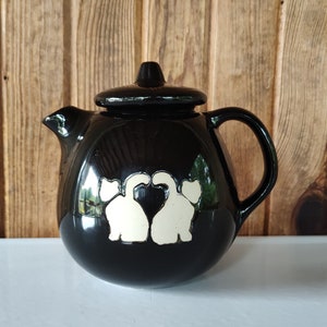 May include: Black ceramic teapot with a white design of two cats facing each other with their tails intertwined. The cats are outlined in white and have white hearts on their chests.