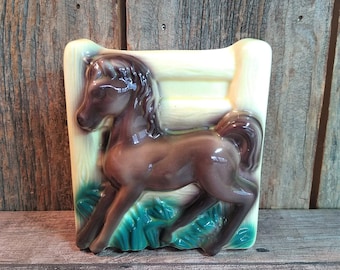 May include: A brown ceramic wall planter shaped like a horse jumping over a fence. The planter has green and yellow accents and is perfect for adding a touch of rustic charm to any home.