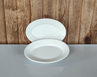 May include: Two white oval-shaped plates are stacked on a gray surface. The plates have a simple, elegant design with a slightly raised rim. The background is a wooden wall with vertical planks.
