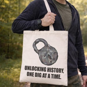 May include: A cream tote bag with a detailed illustration of an antique padlock. The bag has the text "UNLOCKING HISTORY, ONE DIG AT A TIME." The bag is held by a person wearing a dark blue hoodie.
