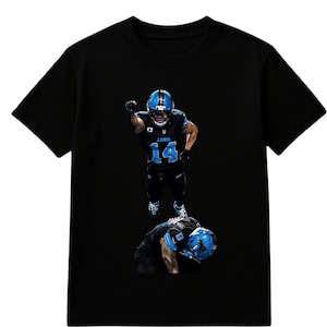 May include: Black t-shirt featuring a graphic of a football player in a blue helmet and uniform, with the number 14. The player is in action, with another player on the ground below.