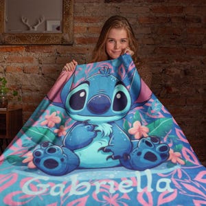 May include: A soft fleece blanket featuring a large blue Stitch character illustration surrounded by pink tropical flowers and green leaves on a blue background, with the name Gabriella printed in white letters at the bottom of the design.