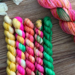 May include: A collection of six twisted yarn skeins featuring a vibrant palette of neon pink, bright orange, sunny yellow, and vivid green. One large skein is positioned diagonally, while five smaller, uniform skeins are arranged vertically.