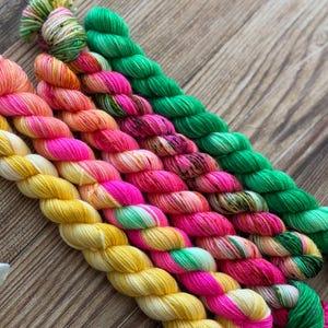 May include: A collection of six twisted skeins of yarn arranged in a diagonal row. The yarn features vibrant, multi-colored hand-dyed patterns including bright yellow, neon pink, orange, and vivid green hues with subtle speckled color variations.