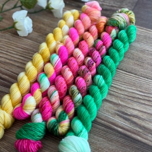 May include: A collection of small, twisted skeins of yarn arranged in a diagonal row. The colors include bright yellow, vibrant pink, orange, and neon green, with some skeins featuring variegated patterns of mixed hues and speckled textures.