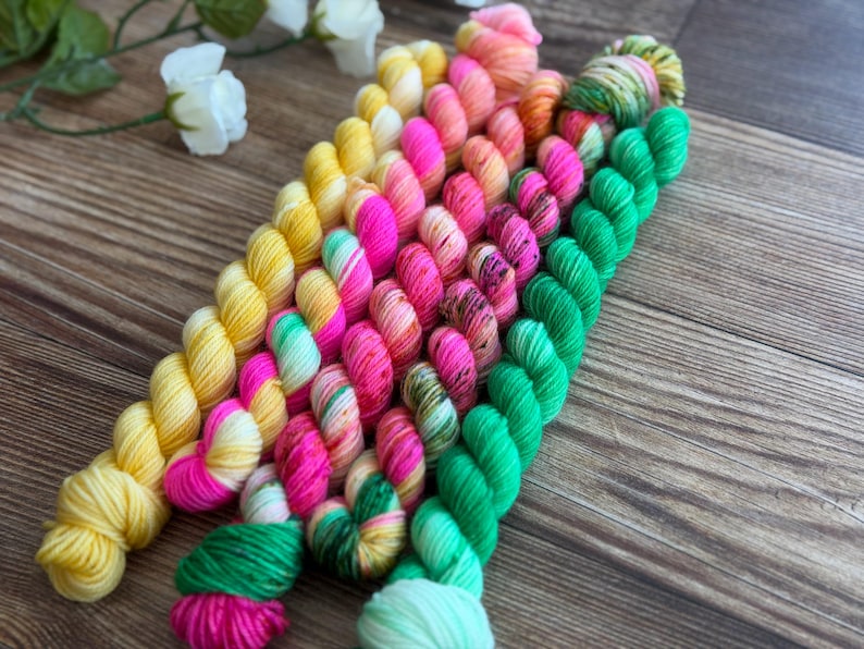 May include: A collection of small, twisted skeins of yarn arranged in a diagonal row. The colors include bright yellow, vibrant pink, orange, and neon green, with some skeins featuring variegated patterns of mixed hues and speckled textures.