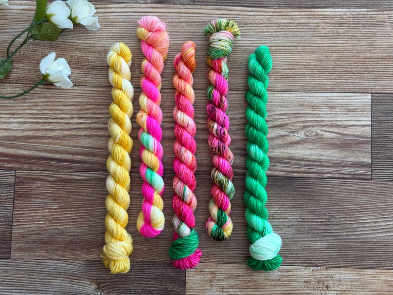 May include: Five twisted skeins of yarn arranged vertically. The first is solid yellow, the second features pink, orange, and light green, the third is pink and orange, the fourth is a variegated mix of pink, green, and white, and the fifth is solid green.
