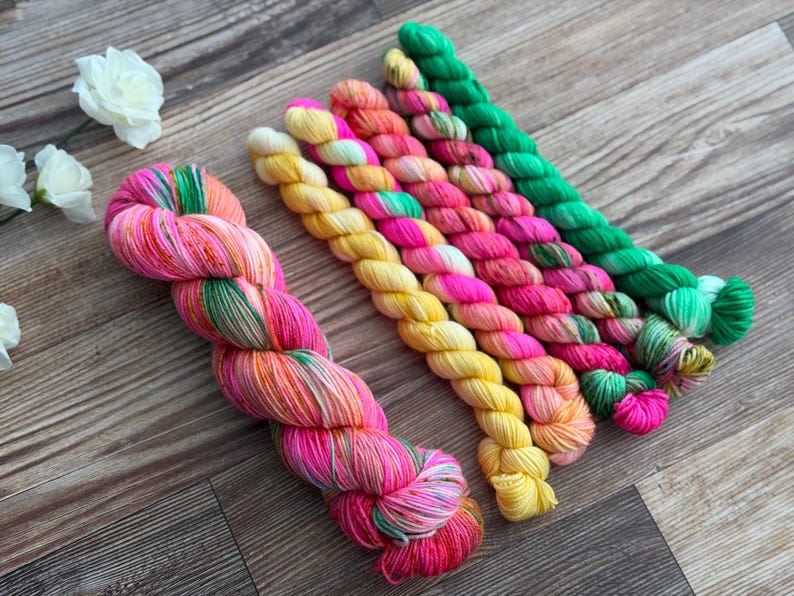 May include: A large skein of variegated yarn in shades of pink, orange, and green, accompanied by five smaller mini-skeins in solid and variegated colors including yellow, bright pink, orange, and green, arranged in a parallel row.