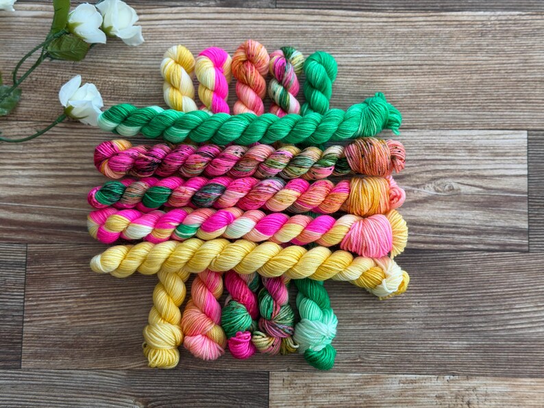May include: A collection of hand-dyed yarn mini skeins arranged in horizontal rows. The yarn features a vibrant palette of neon pink, bright yellow, emerald green, and variegated multi-colored strands, showcasing a twisted texture throughout.