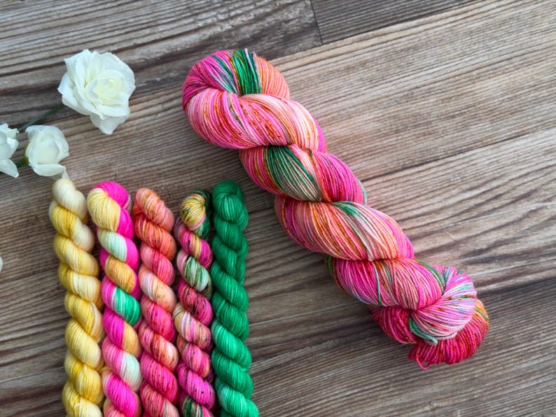 May include: A collection of six twisted yarn skeins featuring a vibrant palette of neon pink, bright orange, sunny yellow, and vivid green. One large skein is positioned diagonally, while five smaller, uniform skeins are arranged vertically.