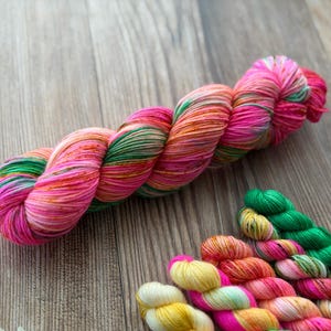 May include: A large skein of yarn featuring a variegated color palette of bright pink, orange, and green, accompanied by several smaller mini-skeins in matching shades of yellow, orange, pink, and green arranged in a row below the main skein.
