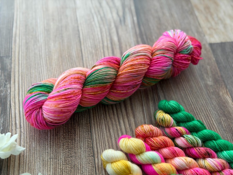 May include: A large skein of yarn featuring a variegated color palette of bright pink, orange, and green, accompanied by several smaller mini-skeins in matching shades of yellow, orange, pink, and green arranged in a row below the main skein.