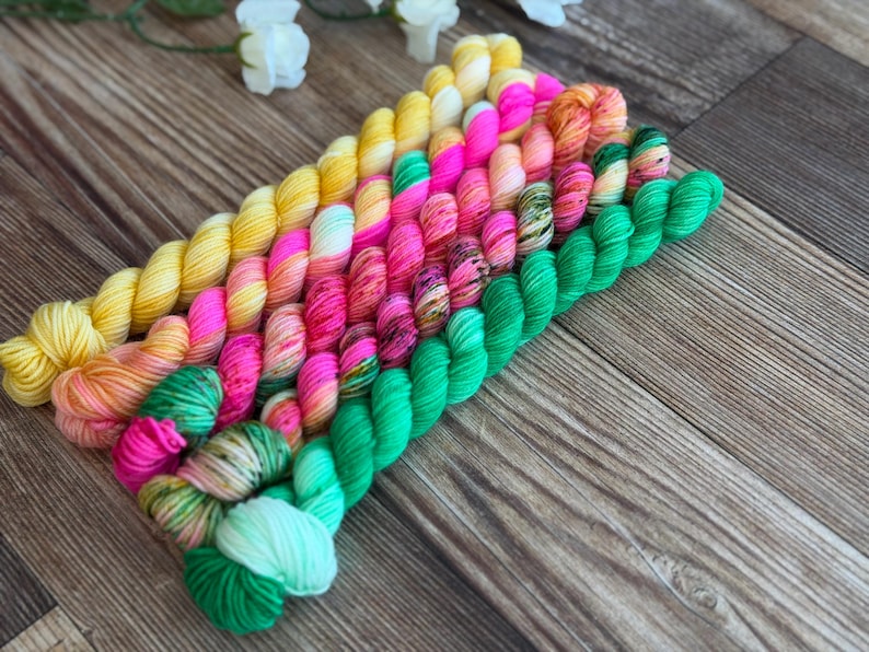 May include: A collection of ten small, twisted skeins of yarn arranged in a diagonal row. The colors transition from bright yellow at the top left to vibrant pink, speckled orange, and finally to various shades of green at the bottom right.