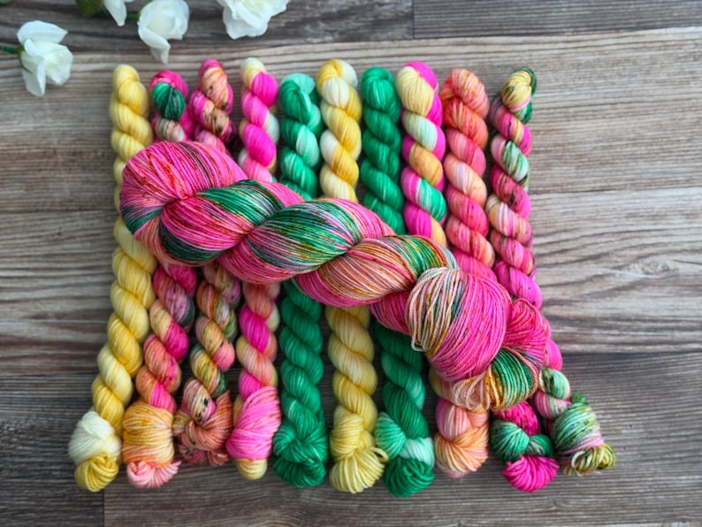 May include: A collection of ten mini skeins of yarn arranged in a row, featuring vibrant colors including bright yellow, hot pink, emerald green, and variegated orange. A single larger, twisted skein of yarn in matching pink and green tones lies horizontally across the top of the vertical row.