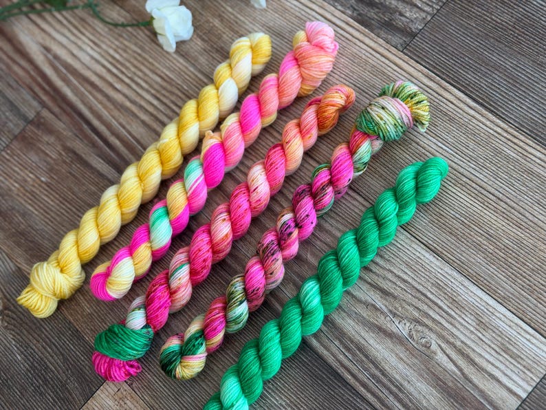 May include: Five twisted skeins of yarn arranged diagonally. The first is solid yellow, the second is a blend of pink and yellow, the third is a mix of pink and orange, the fourth is a variegated blend of pink, green, and yellow, and the fifth is solid green.