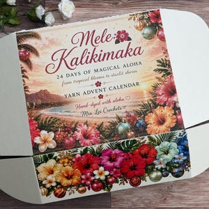 Könnte beinhalten: Ein weißer Karton mit einer tropischen Sonnenuntergangsszene, Hibiskusblüten, Palmen und Weihnachtskugeln. Der Text lautet Mele Kalikimaka, 24 Days of Magical Aloha, Yarn Advent Calendar, Hand-dyed with aloha, Mrs. Lei Crochets.