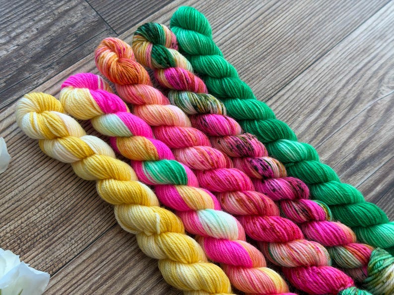 May include: Five skeins of hand-dyed yarn arranged diagonally. The colors include a solid bright yellow, a vibrant hot pink, a soft peach and orange blend, a variegated pink and green mix, and a solid vivid green. Each skein is twisted.