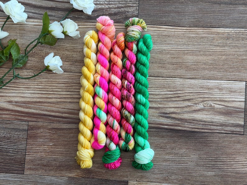 May include: Five twisted skeins of yarn arranged vertically. The colors include solid bright yellow, neon pink with white accents, a variegated blend of orange and pink, a multi-colored mix of green and pink, and a solid vibrant green.