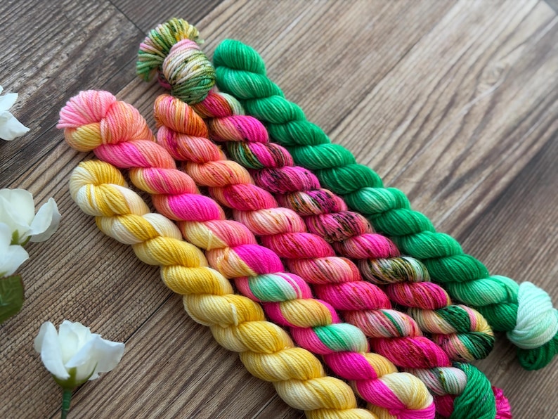 May include: A collection of six twisted skeins of yarn arranged in a diagonal row. The yarn features vibrant, multi-colored hand-dyed patterns including bright yellow, neon pink, orange, and vivid green hues with subtle speckled color variations.