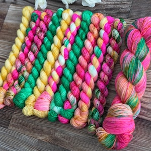 May include: A collection of ten twisted skeins of yarn arranged in a row. The yarn features a vibrant color palette including bright yellow, hot pink, emerald green, and orange, with some skeins displaying variegated patterns of these mixed colors.